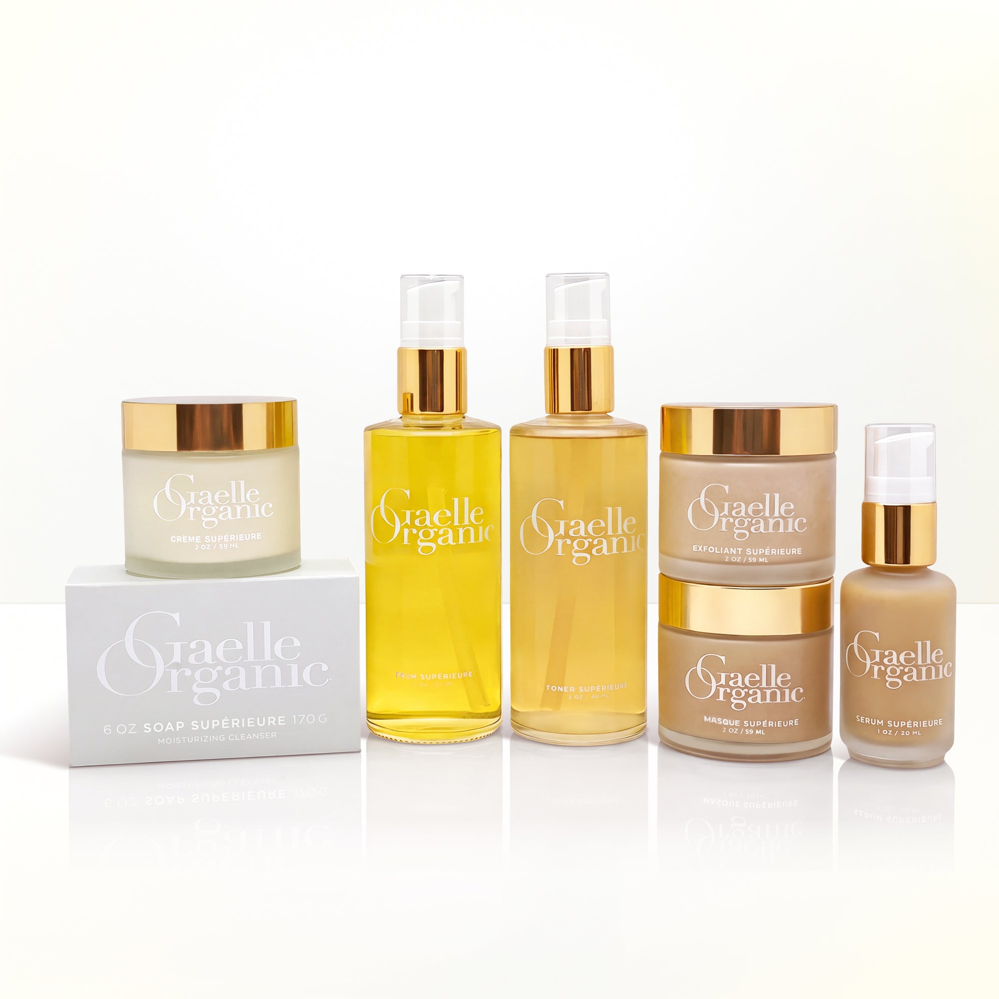 A set of five Graelle Organic skincare products arranged on a white reflective surface, including a jar, two spray bottles, two cream jars, and a small bottle, with a white box. The products feature gold caps and metallic gold lettering, with the creams and oils in shades of beige, yellow, and light brown.