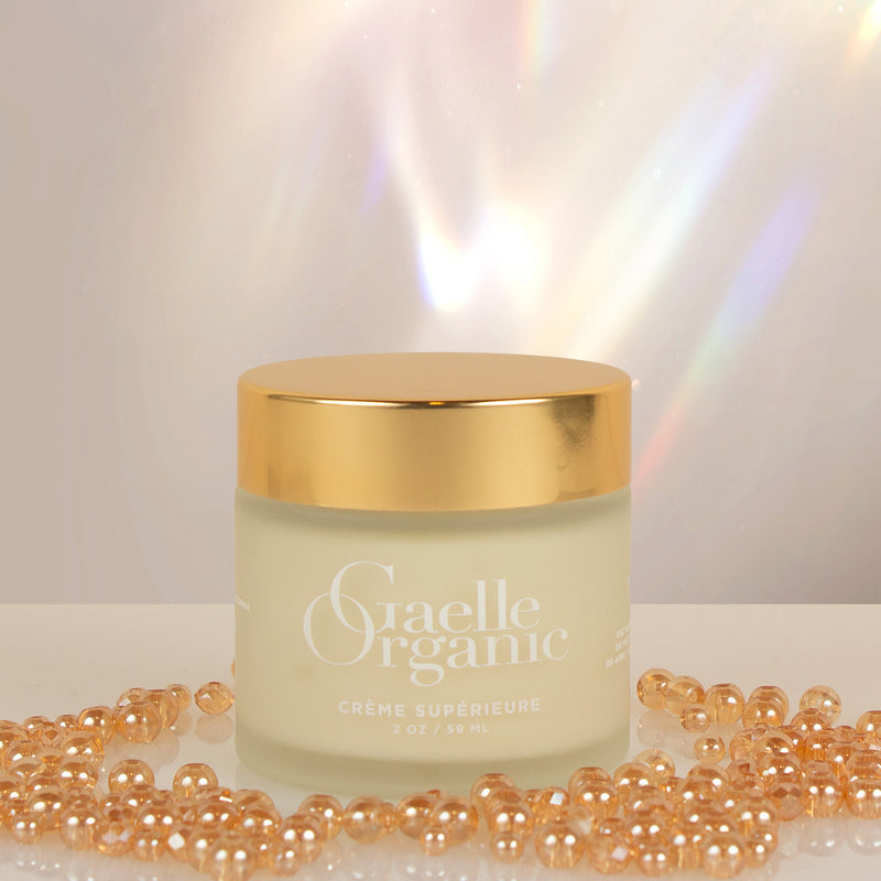 Creme Superiure, an organic moisturizer for aging and dry skin, on a white reflective surface with golden beads.